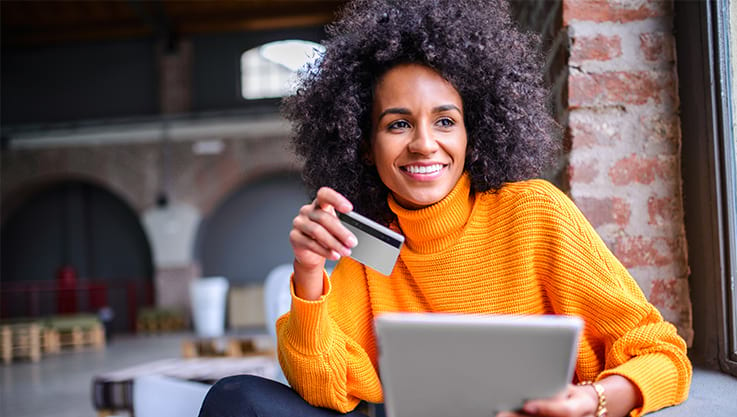 A black woman with an orange sweater holds a tablet and credit card.