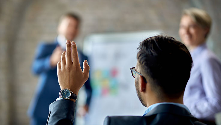 A person presenting at the front of the room calls on someone in the audience raising their hand. 