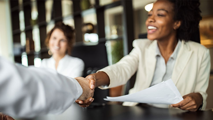 Two professionals sit at a desk with papers, interviewing a person facing them. 