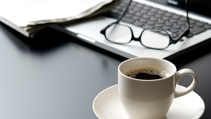 On a dark colored desk sits a silver laptop, glasses and a cup of coffee on a matching saucer.