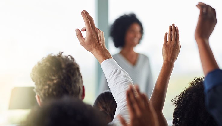 A businessperson stands in front of a group of collegues raising their hands. 