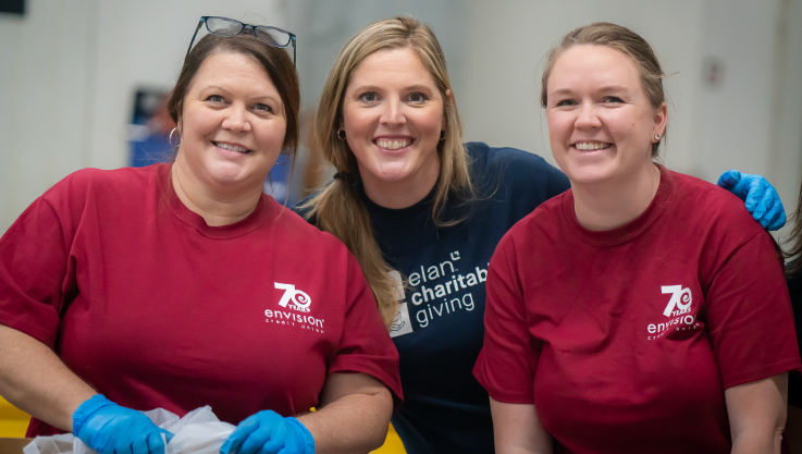 Three women smile at the camera while packaging food at a food shelf.