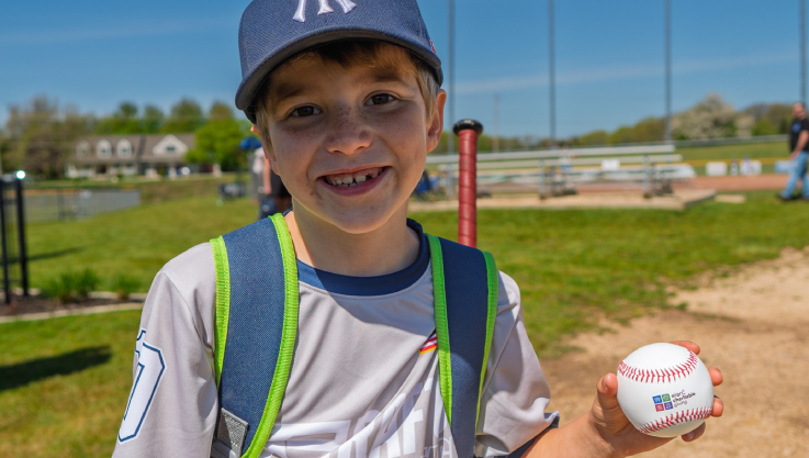 A child stands outside holding a baseball with the Elan Charitable Giving logo. 