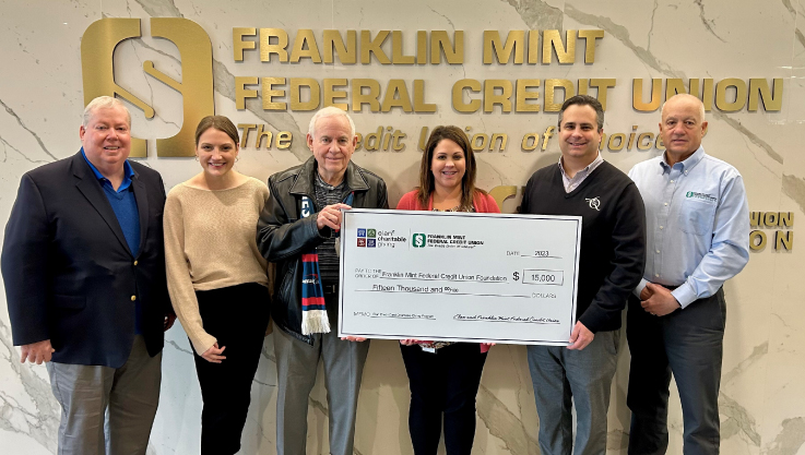 Six adults hold a large check in front of a marble wall with gold text that reads "Franklin Mint Federal Credit Union."