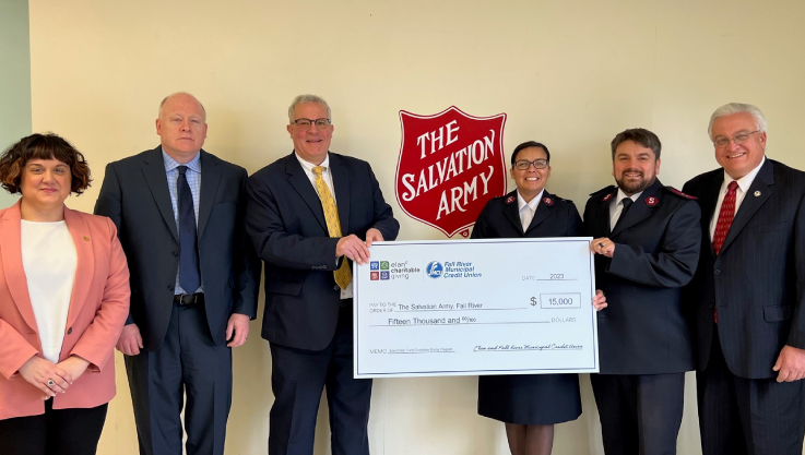 Six adults hold a large check in a build lobby. Centered above the check is The Salvation Army's logo. 