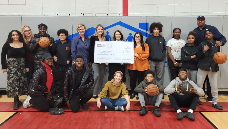 A group of adults and teenagers stand and sit at the end of a basketball court. Three adults in the center are holding a large check. 