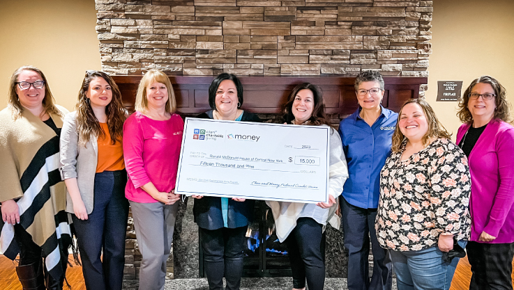 Eight adults stand in front of a fireplace surround. The three in the center hold a large check.