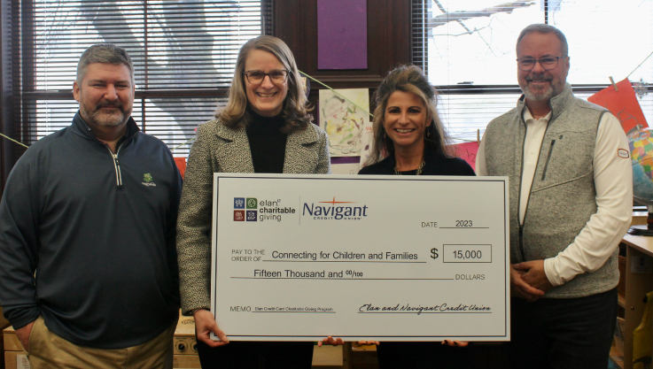 Four adults hold a large check in front of two windows separated by a purple wall. 
