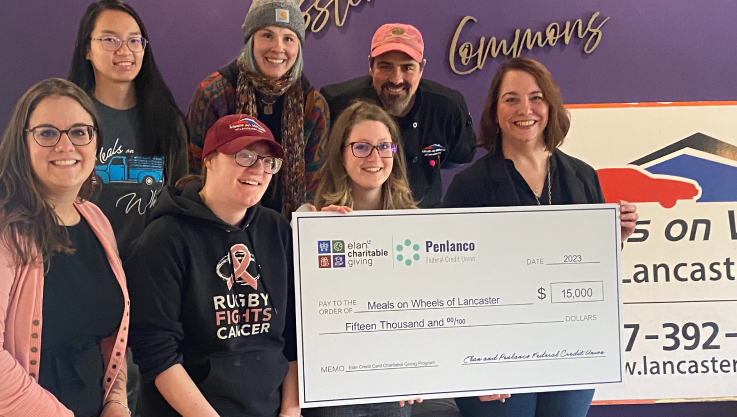 Seven adults pose for a photo holding a large check. They are standing in front of a purple wall. 