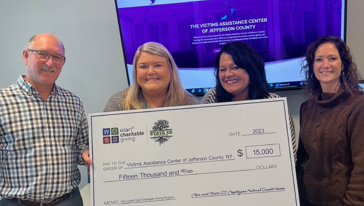 Four adults stand in front of a monitor in a conference room holding a large check. 