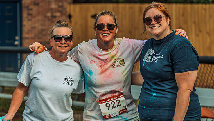 3 women linking arms smile at the camera. The middle is wearing a running bib and has a shirt full of color.