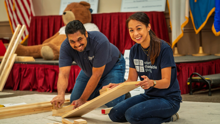 Two Elan volunteers look at the camera smiling while assembling a bed frame. 