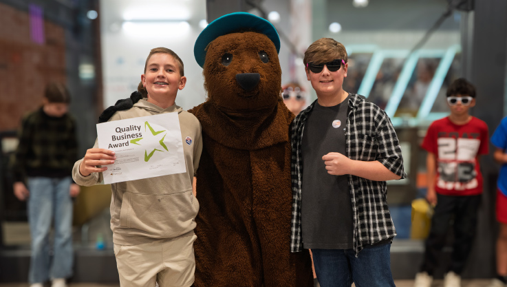 Two children stand with a mascot smiling at the camera. The student on the left is holding an award certificate. 