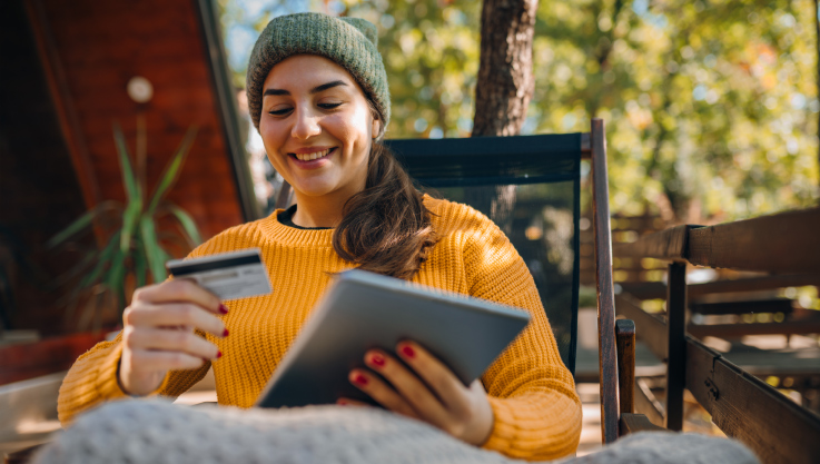 A woman sits outside at a cabin holding a tablet and credit card.