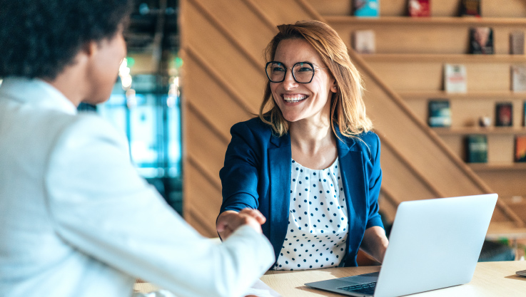A woman sits at a table with her laptop, she is shaking the hand of someone across the table. 