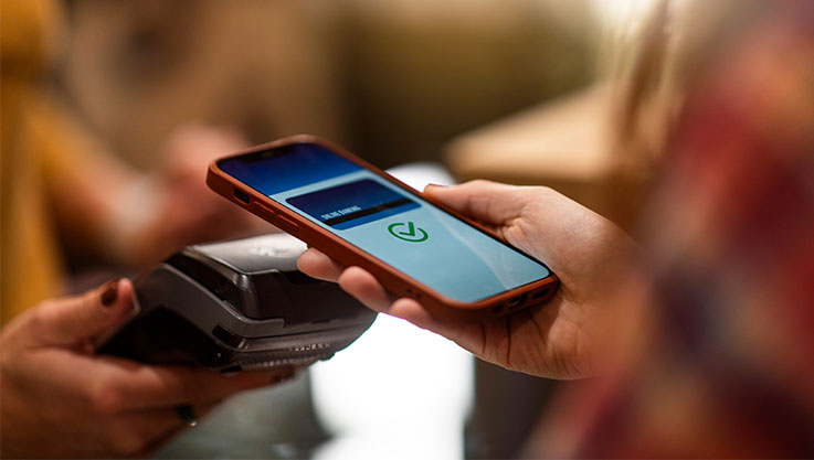 A person holds their smartphone over a credit card terminal. The phone screen shows a picture of a credit card and a green check mark. 