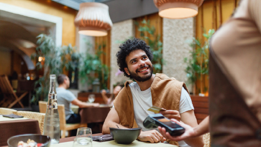 A man sits at a table inside a restaurant. He holds a credit card over a payment terminal held by the server. 